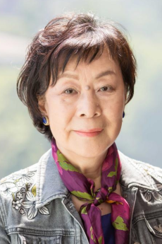 An older Japanese woman with short hair wears earrings and a neck scarf. She wears a blue shirt underneath a grey denim jacket.